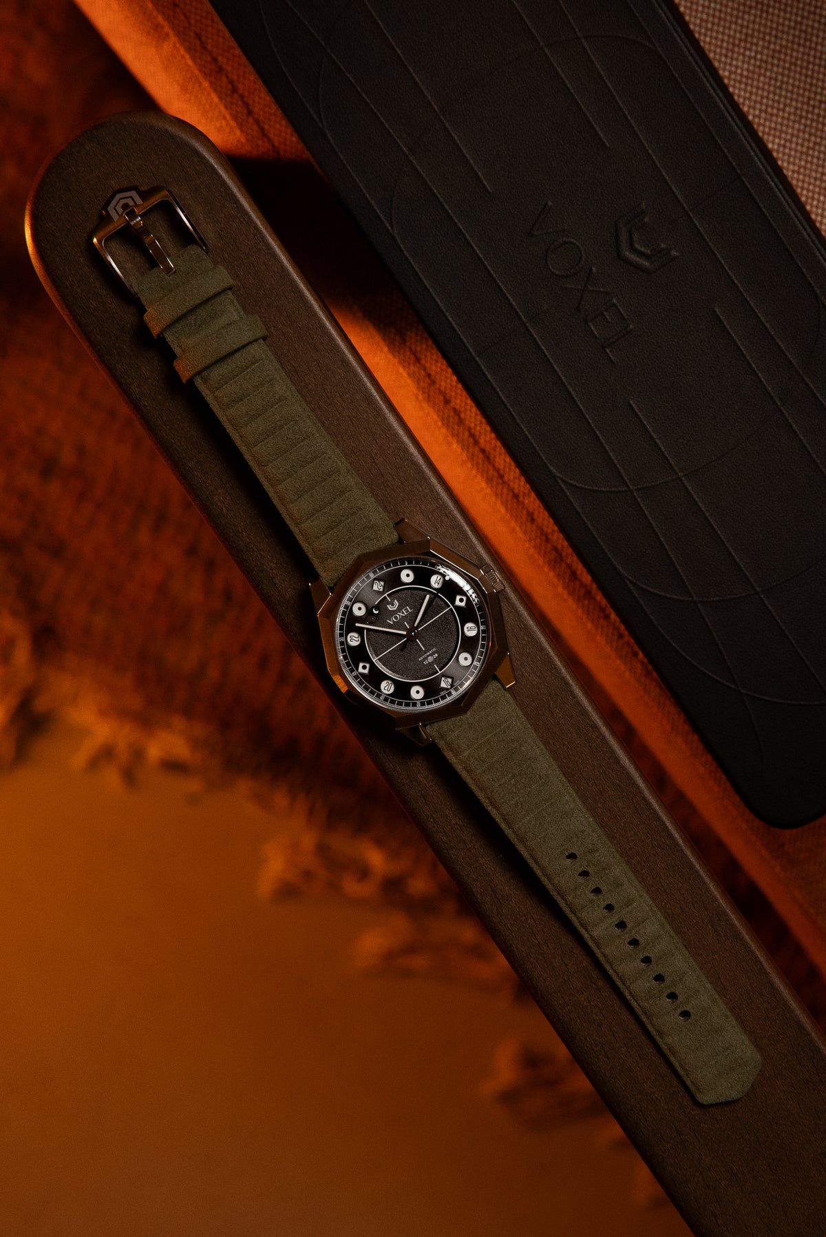 VOXEL watch with dark dial and olive green canvas strap, resting beside its black presentation box in warm light