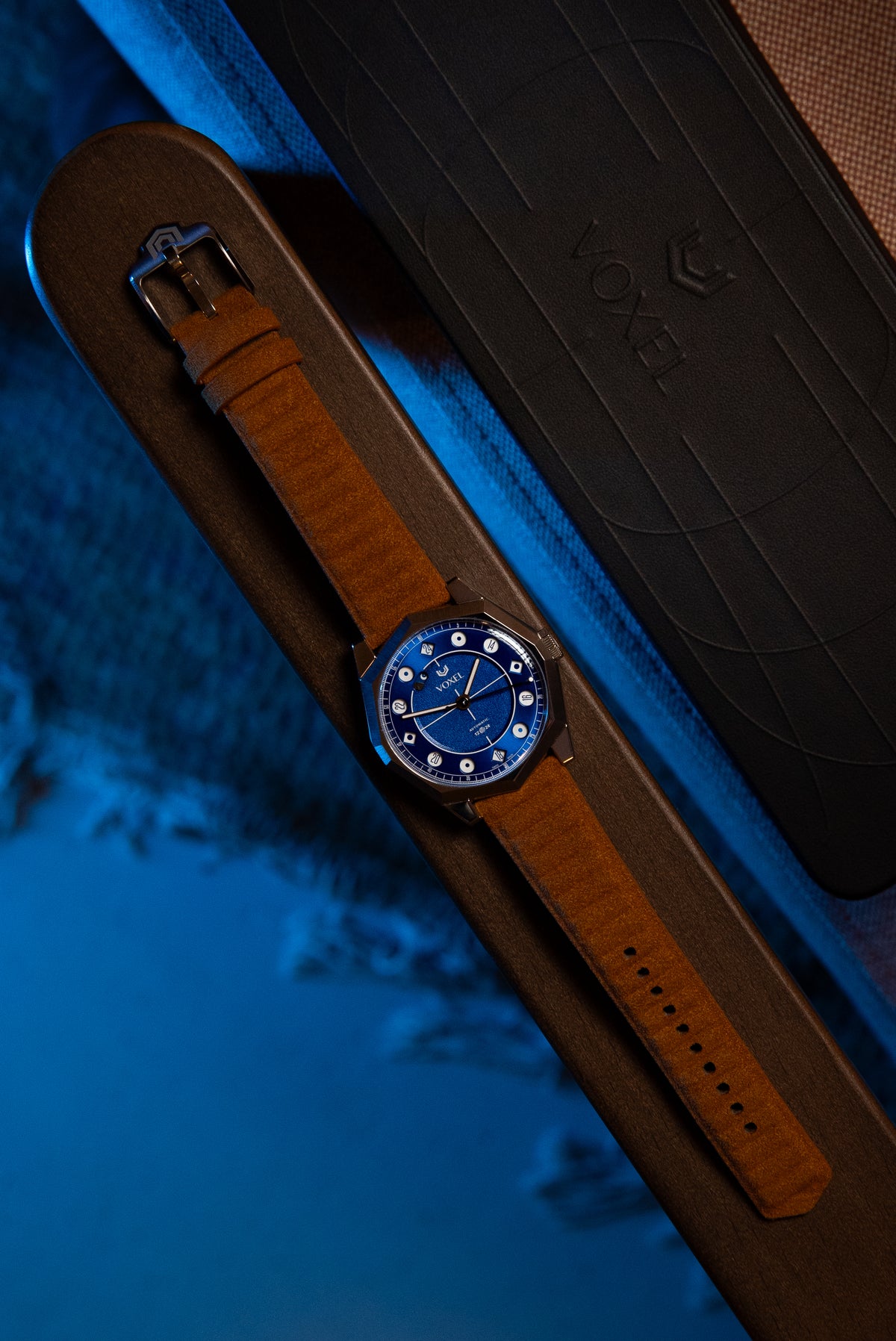 VOXEL watch with blue dial and tan suede strap, resting beside its black presentation box in cool blue light
